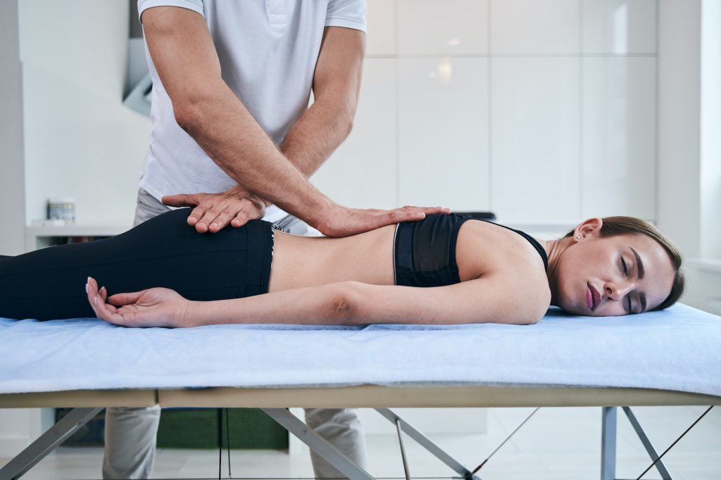 Unrecognized man osteopath stretching back of beautiful lady in rehabilitation clinic